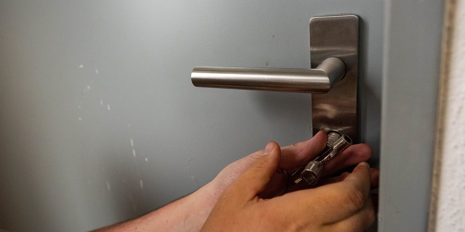 Close-up of two hands fitting a cylinder lock below the steel handle of a grey door