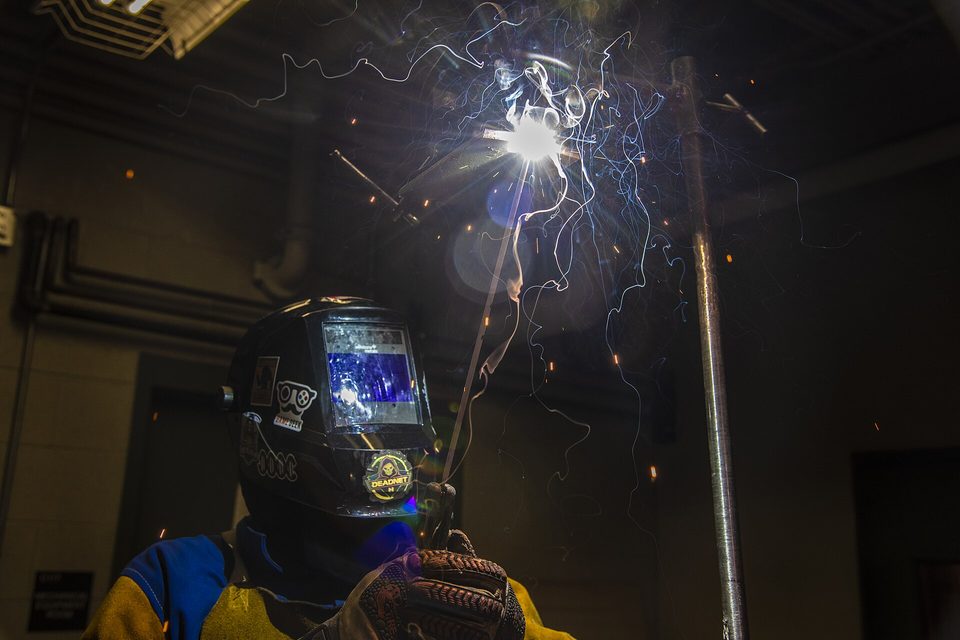 A welder in a dark helmet strikes an arc, throwing bright sparks in a dim shop