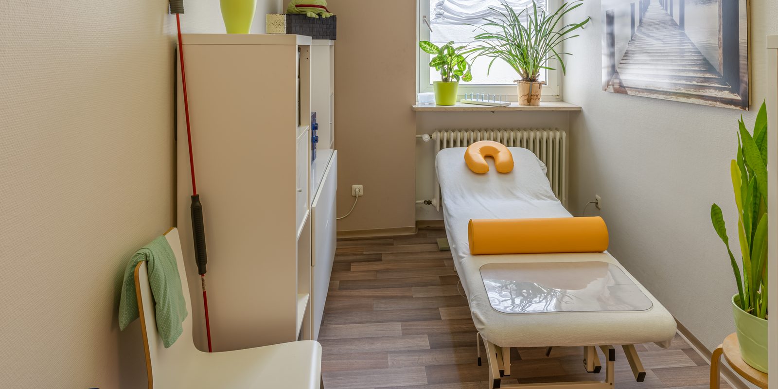 A small treatment room with a massage table, orange bolster and face cushion, houseplants and a sunny window