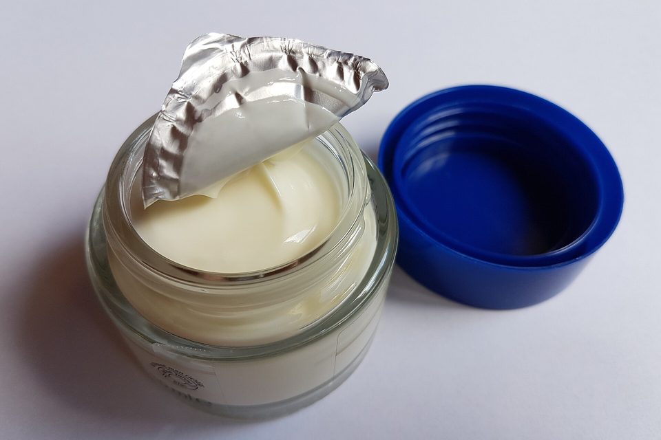 An open glass jar of pale cream with its foil seal peeled back and a blue lid resting beside it