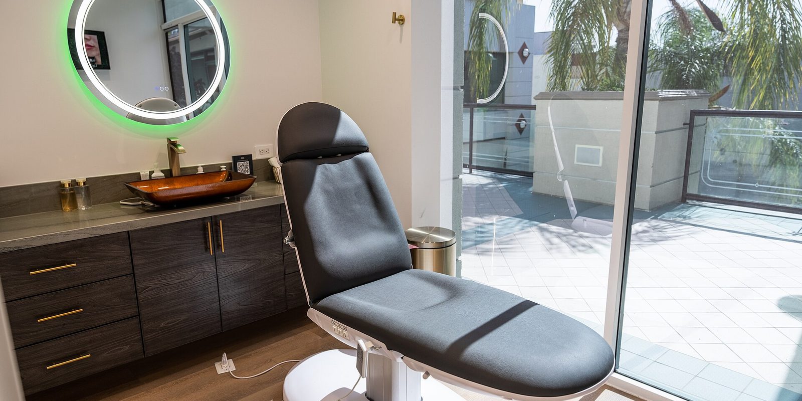 A treatment room with a reclining grey chair, vanity sink and round backlit mirror beside a glass wall