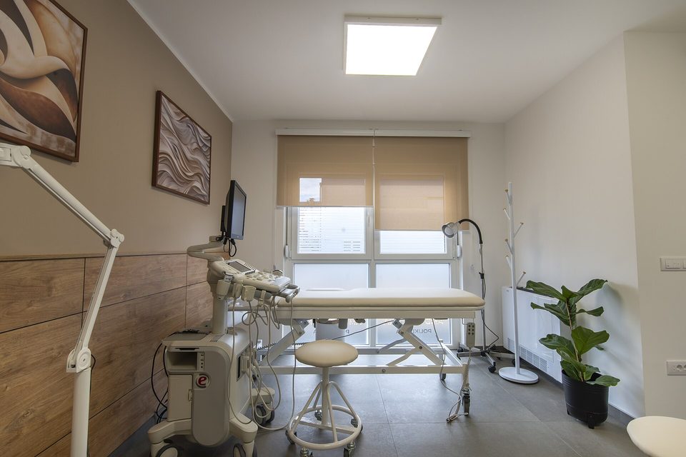 An empty clinic exam room with an ultrasound cart, padded exam table, wheeled stool and examination lamp
