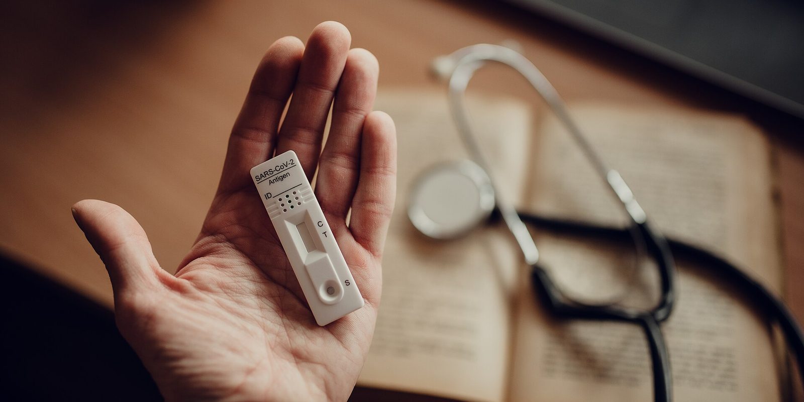 An open palm holding a rapid antigen test cassette, with a stethoscope resting on an open book behind it
