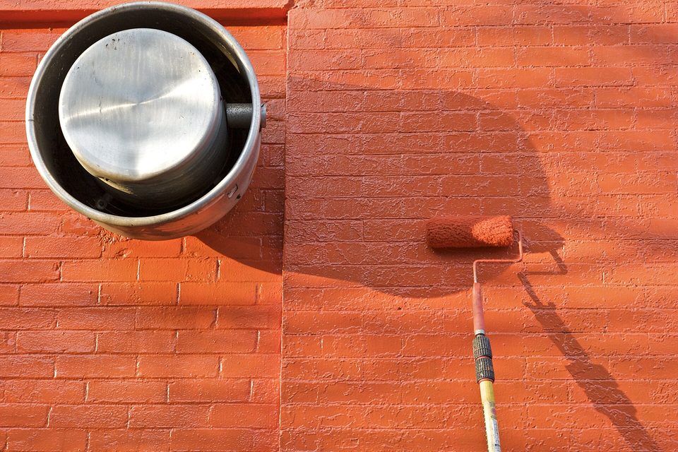 A roller on an extension pole spreading orange paint across a brick wall beside a metal wall vent