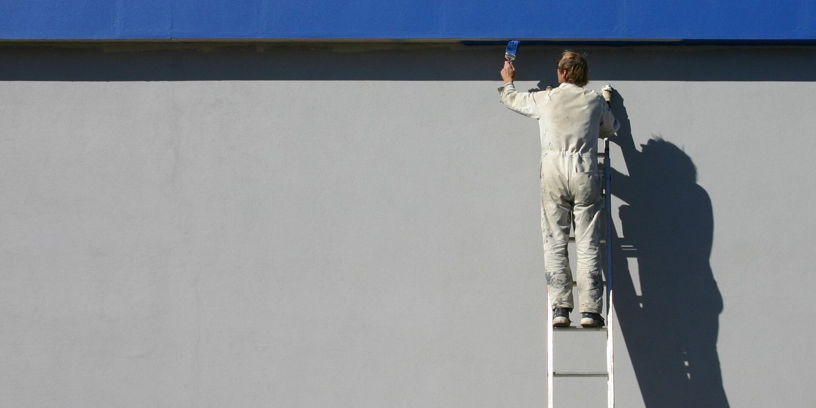 A painter in white overalls on a ladder, brushing a blue stripe along the top of a grey exterior wall