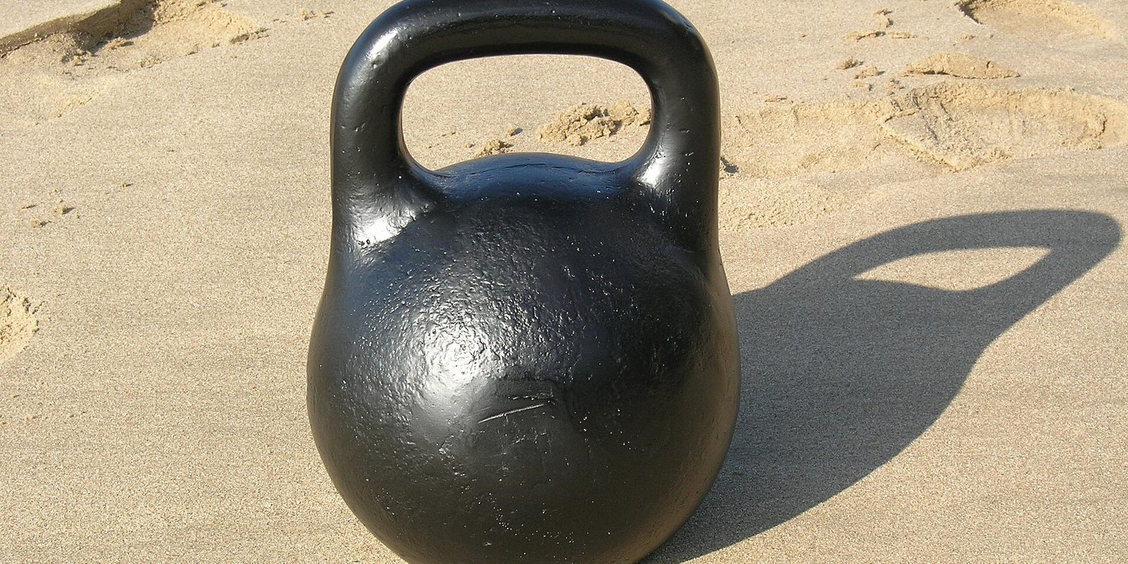 A black cast iron kettlebell standing on dry sand, casting a long shadow in bright sun