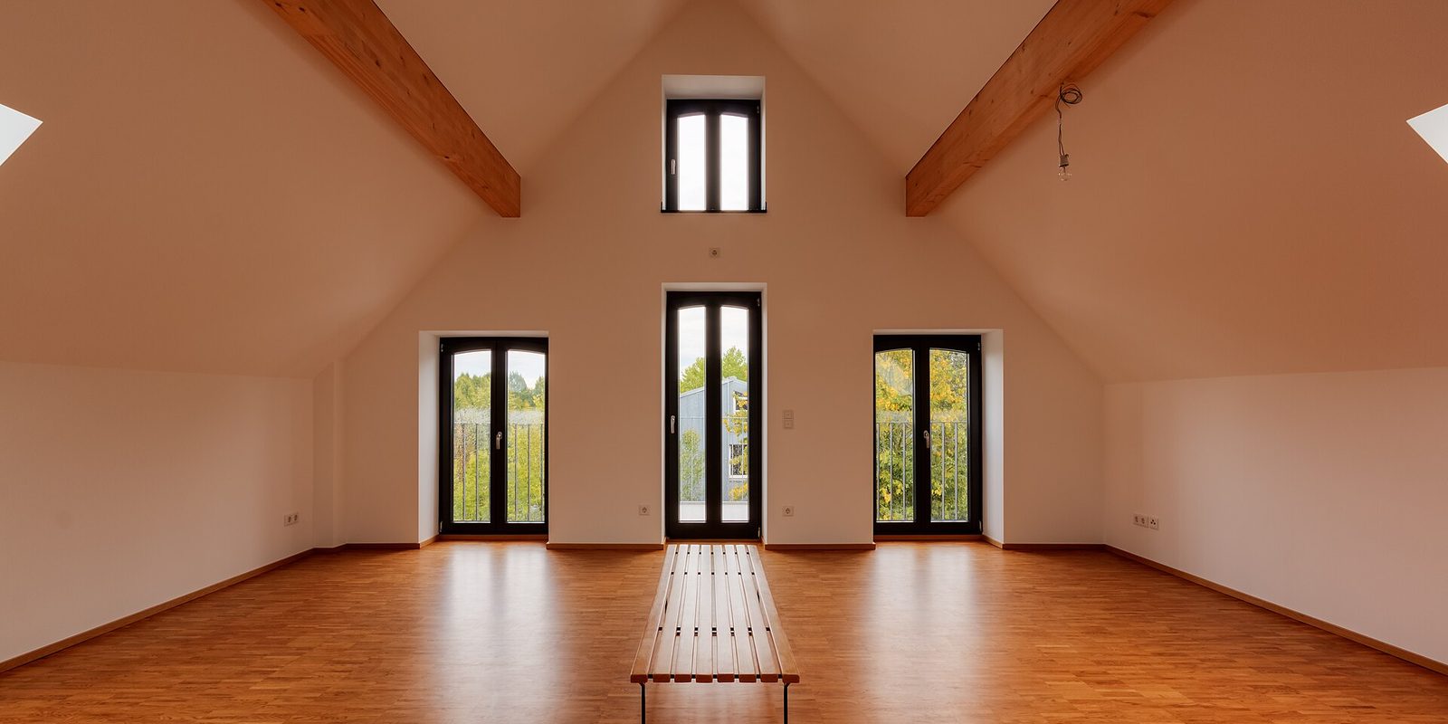 An empty attic room with a vaulted ceiling, wood beams, tall windows and a bare wood floor