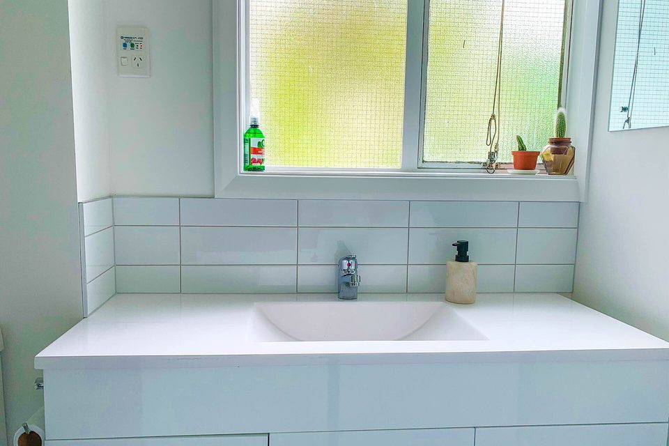 White bathroom vanity with a wide basin, tiled splashback and small potted plants on the windowsill