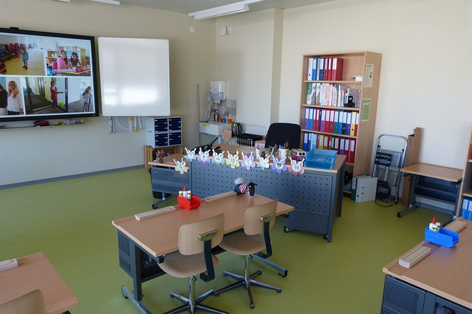 A classroom with a wall mounted display screen, whiteboard, folder shelves and pupil desks
