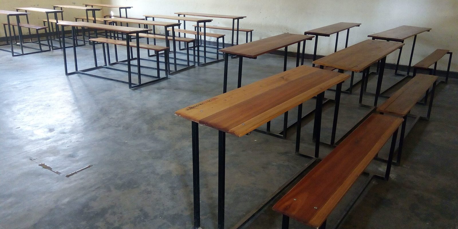 An empty classroom with rows of wooden bench desks on a bare concrete floor