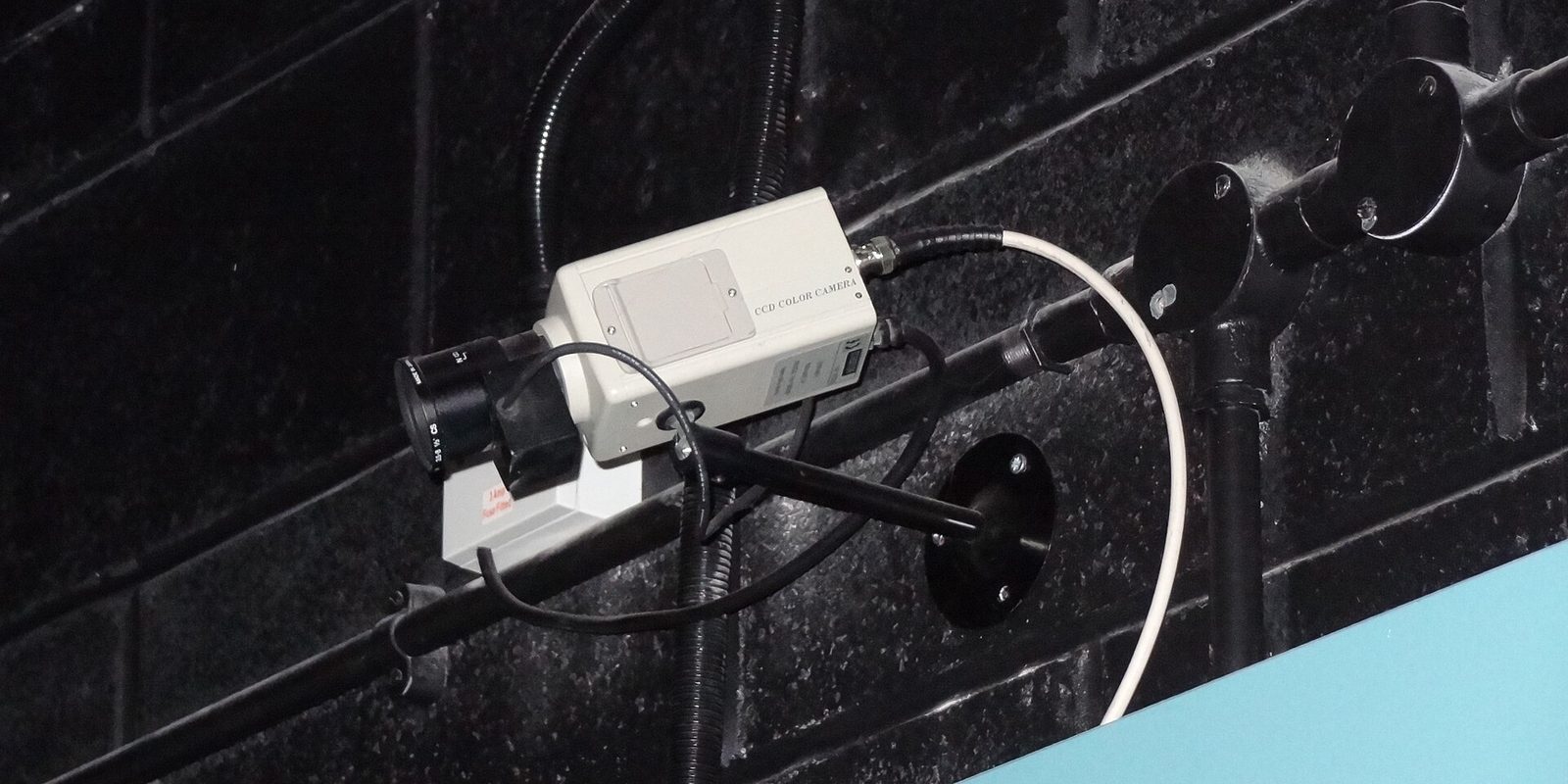 A white boxy CCD surveillance camera bolted to a dark painted brick wall, cables trailing behind it