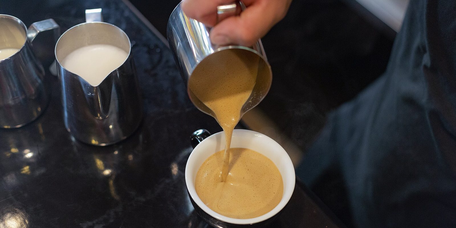 A hand pouring steamed coffee from a steel jug into a white cup, with milk jugs on a dark counter