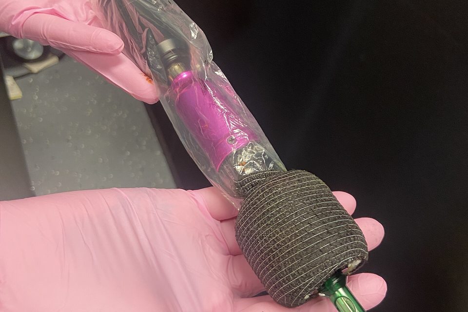 Gloved hands holding a tattoo machine sealed inside a clear plastic barrier bag