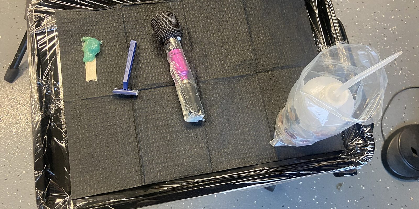 Overhead view of a plastic-wrapped tattoo station tray holding a razor, ointment, bagged machine and squeeze bottle
