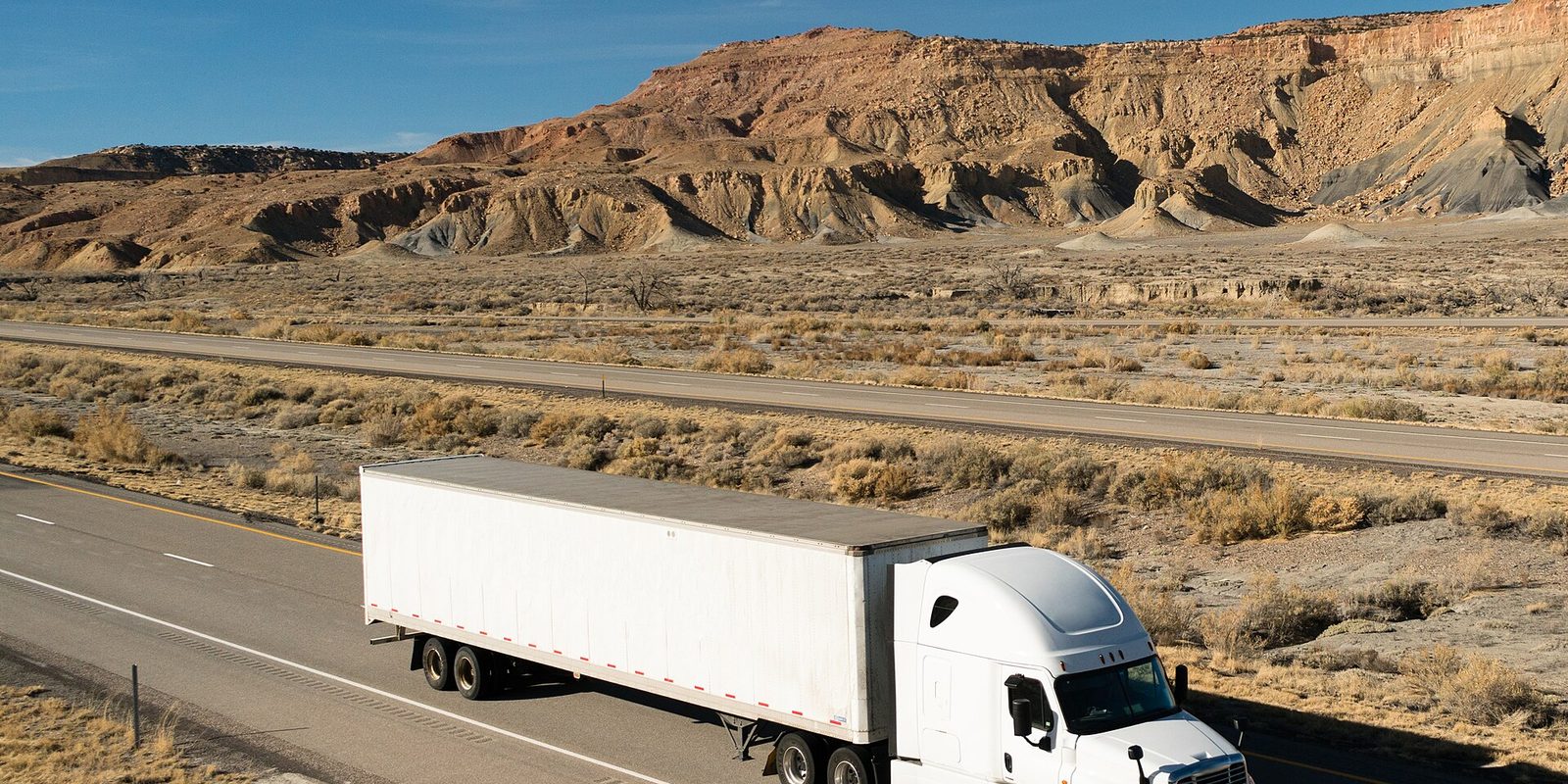 A white semi truck with a dry van trailer on a desert highway below eroded rock cliffs