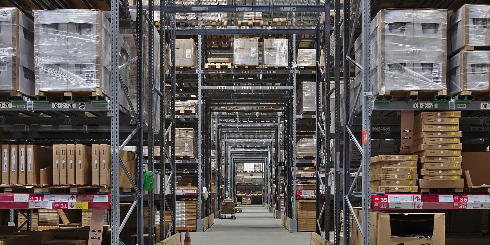 A long warehouse aisle lined with tall racking holding shrink wrapped pallets and labelled cartons