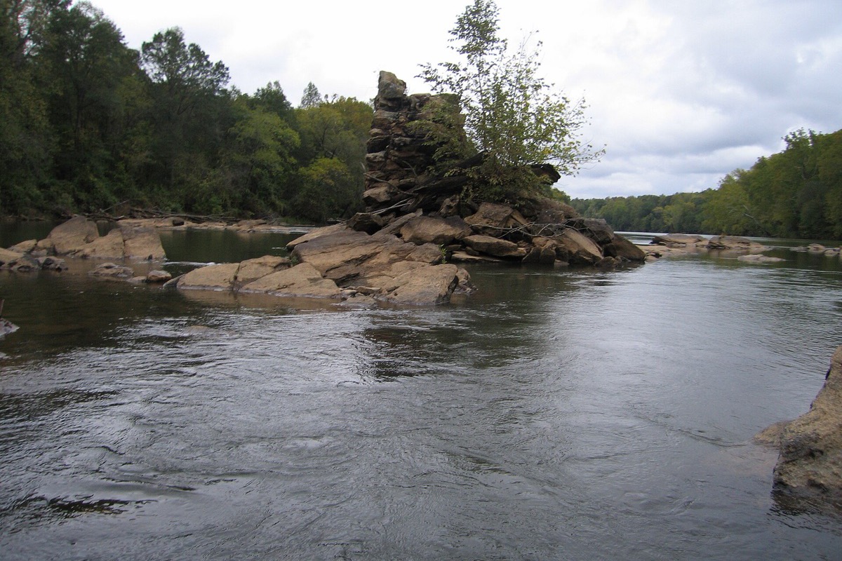 A wide Alabama river running low over pale rock, the stone piers of a long gone covered bridge standing as a small tree topped island in midstream, hardwood forest closing in on both banks.