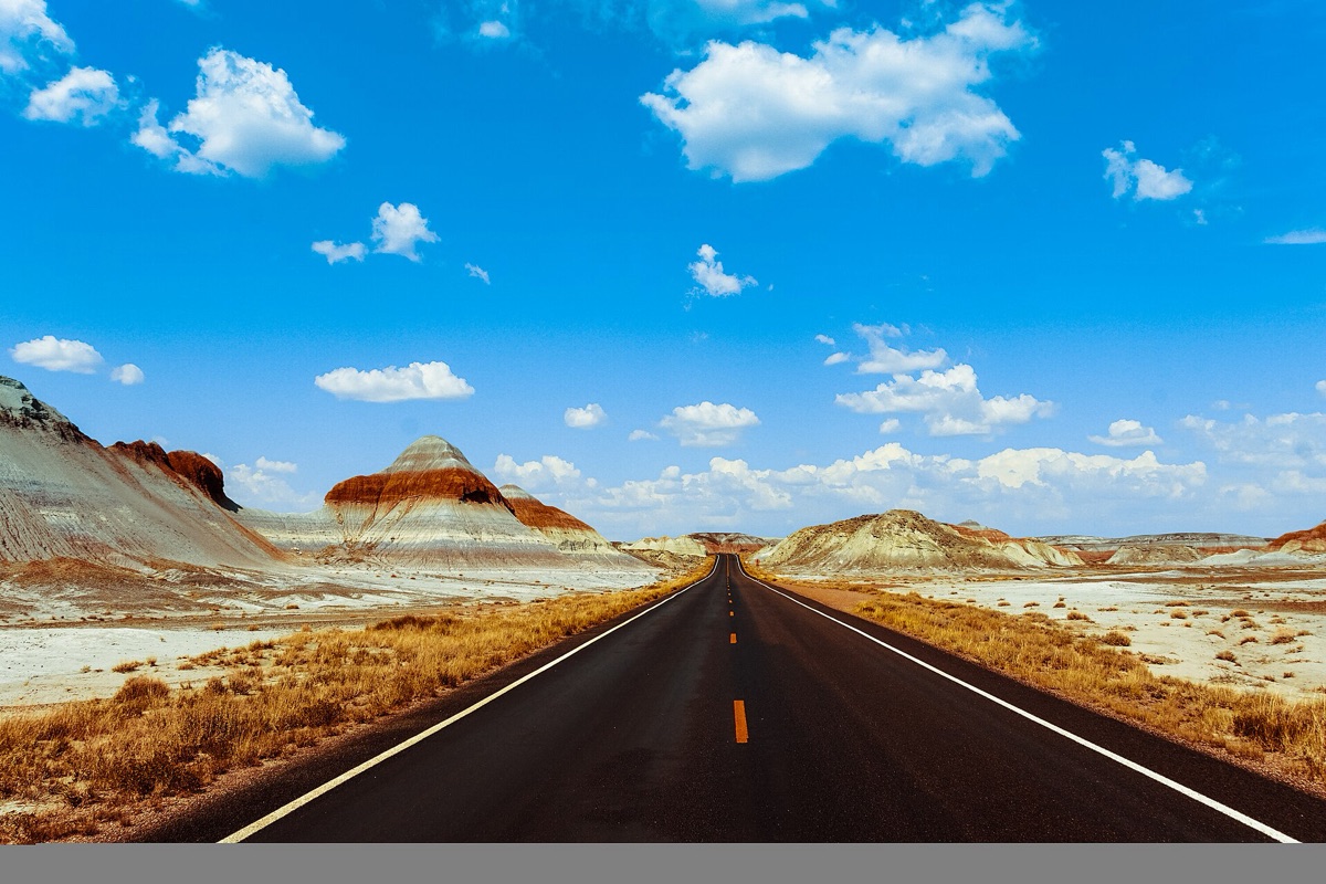 A straight two lane road running away across the Arizona desert between banded red and grey clay hills, dry grass on both shoulders and towering white cloud in a deep blue sky.