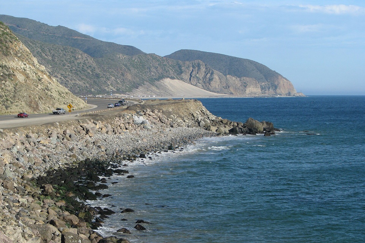 The California coast road cut into a bare headland above the Pacific, traffic strung out along it, a rocky shore and green surf below and the cliffs running away into the haze.