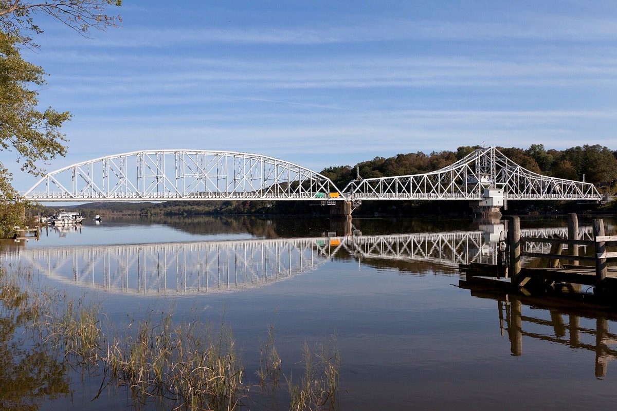 A long white steel truss swing bridge carrying a road low across the Connecticut River, the whole span mirrored in flat calm water, with wooded hills behind it under a clear blue sky.