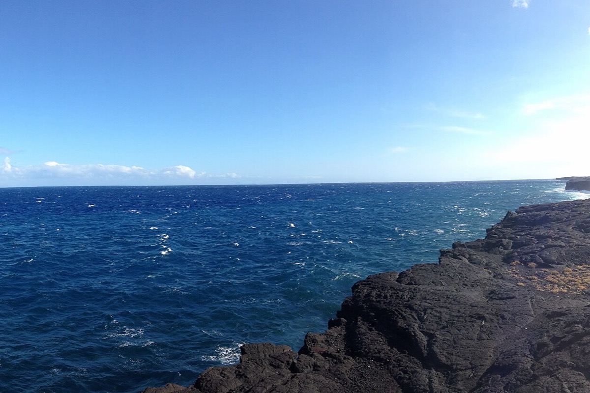 A black shelf of old lava running out into the Pacific in Hawaii, deep blue water breaking white along its edge and a long low horizon under a bright sky.