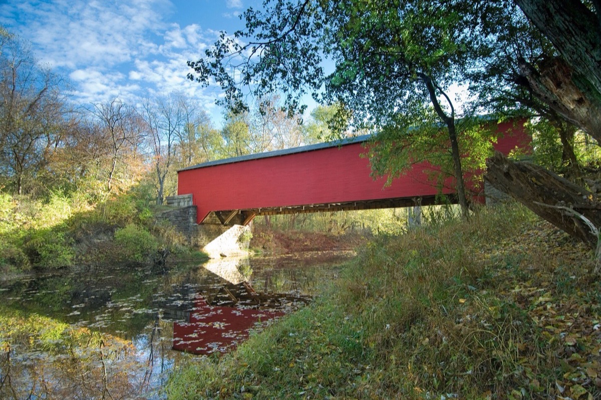 A red painted covered bridge crossing a still Indiana creek in autumn, its reflection broken on the water, and yellow and rust coloured hardwoods crowding both banks.