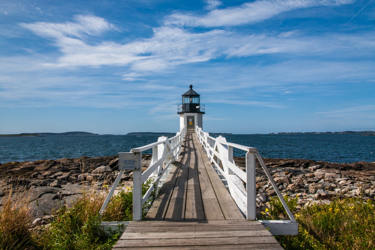 A narrow white railed wooden walkway running out over granite boulders to a small Maine lighthouse, the bay and a low wooded shore behind it under a blue sky.