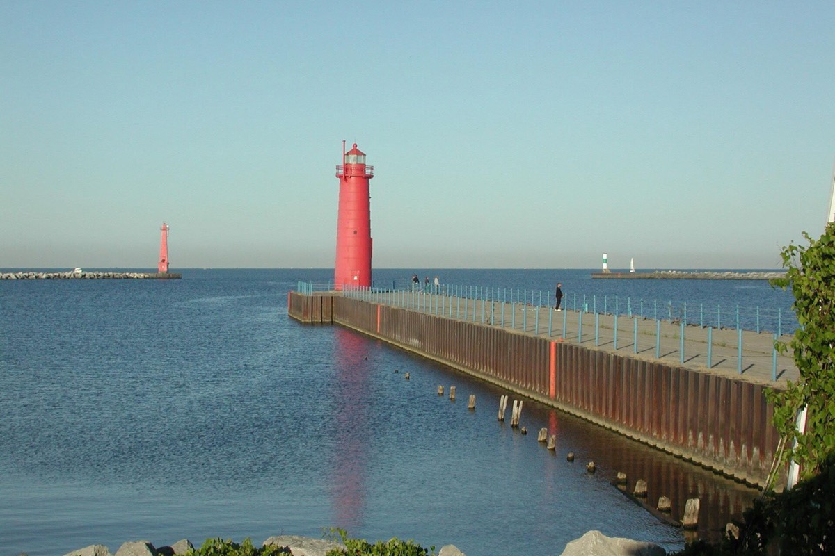 A red steel pierhead light at the end of a long timber pier running out into Lake Michigan, the water flat and pale blue and a second light standing off on the breakwater.
