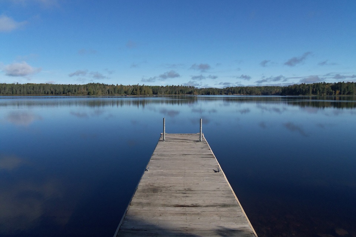 A weathered wooden dock running out into a Minnesota lake so still that the far shore and the whole sky are held in it, dark spruce forest around the entire shoreline.