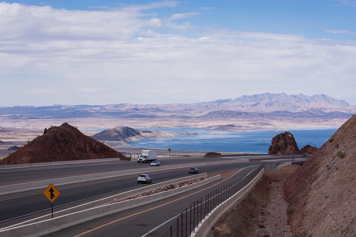 A Nevada road dropping down through dark red rock cuttings toward a wide desert lake, a few vehicles on it and a long bare mountain range standing across the far side of the water.