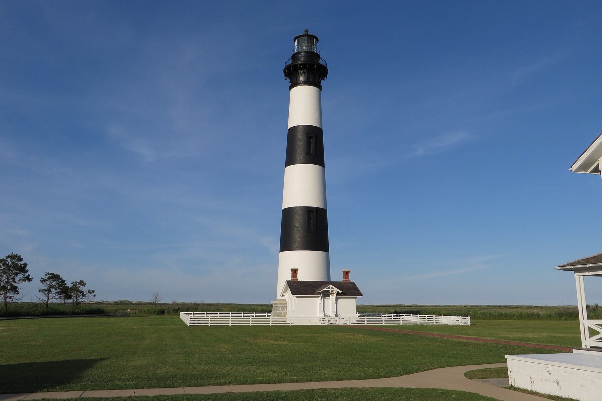 A tall black and white banded North Carolina lighthouse standing over its white keeper's house on mown grass, a white fence around the base and a clear sky behind.