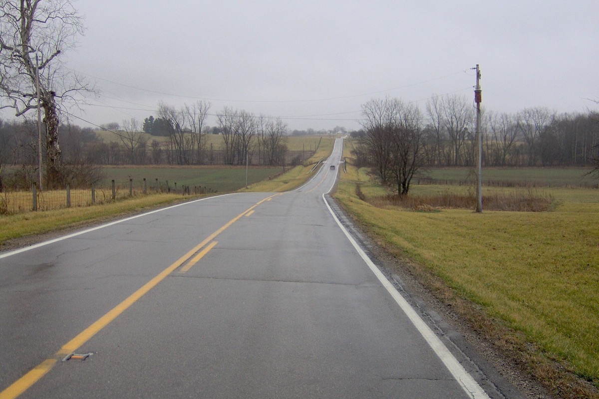 A two lane Ohio road running straight over low rolling farmland on a misty morning, bare trees and a line of poles along the near shoulder and a single vehicle far ahead.