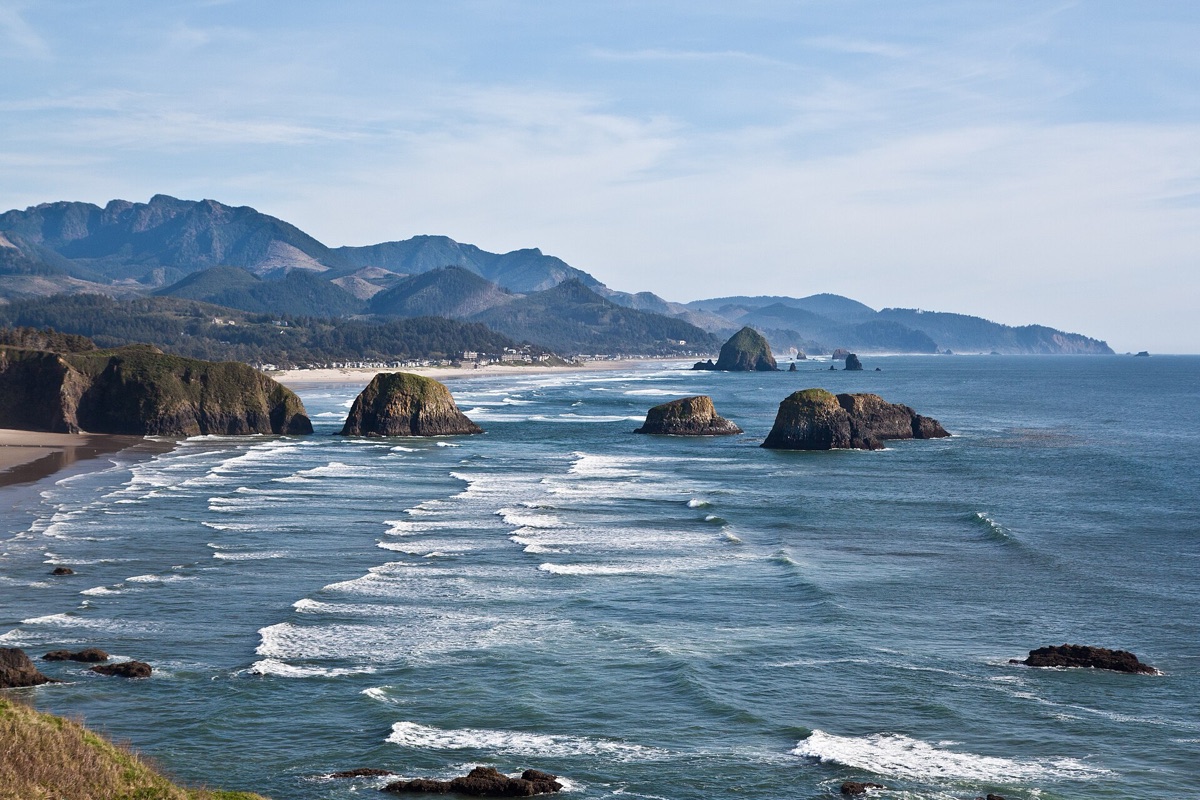 The Oregon coast seen from a headland, long lines of surf breaking over dark sea stacks, a curve of beach running north and forested coastal mountains standing behind it.