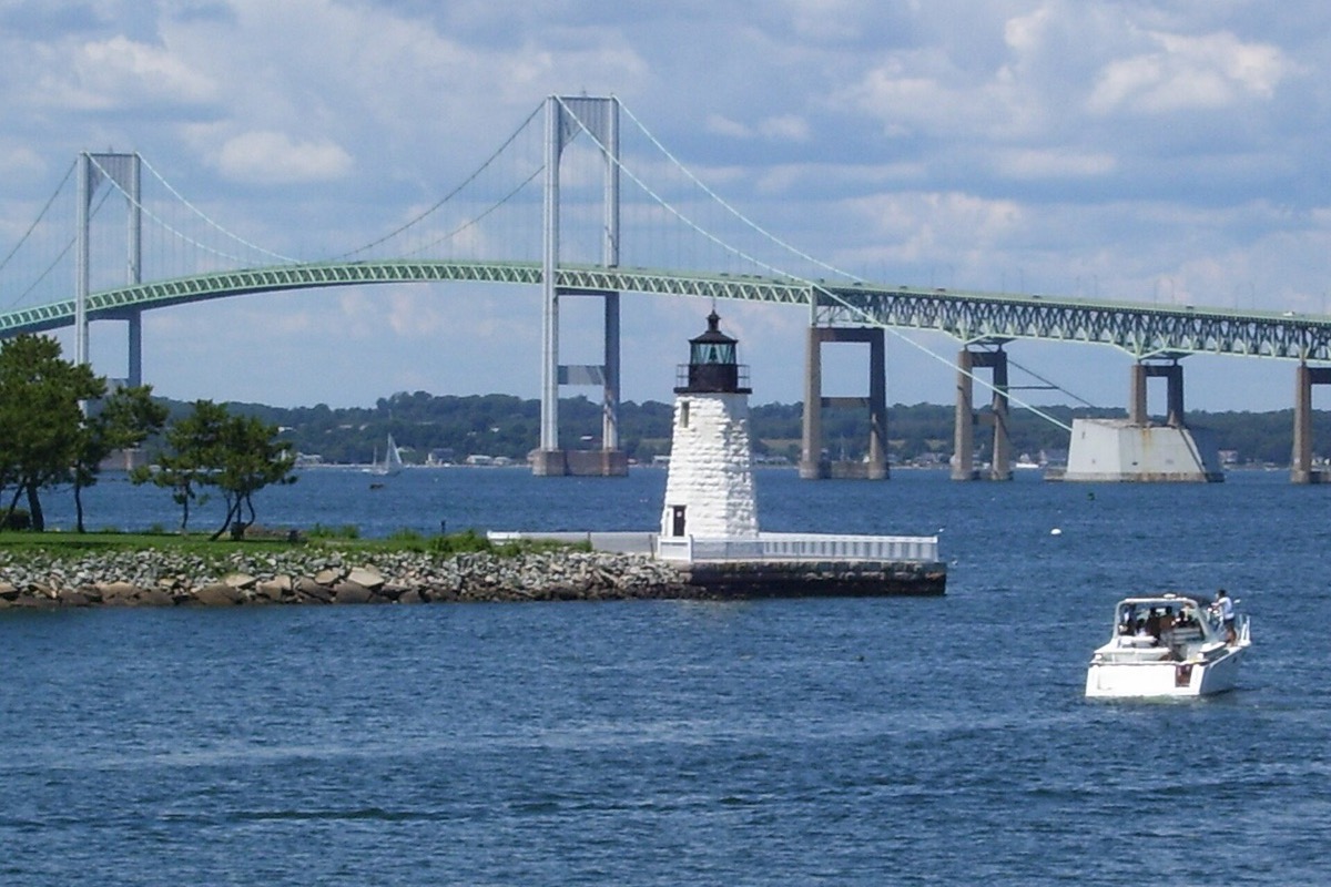A small white Rhode Island harbour light on a stone pier with a motor cruiser passing it, and the long green suspension span across the bay behind under a bright sky.