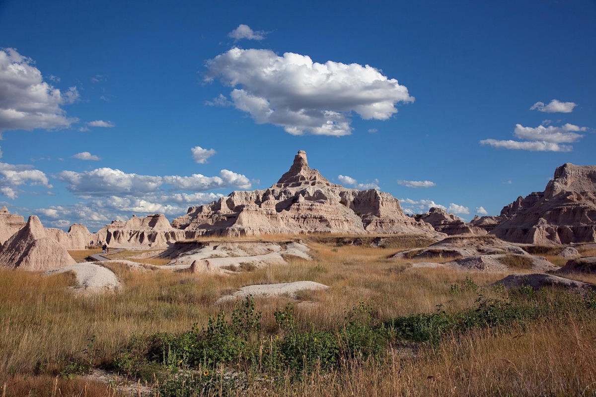 Eroded South Dakota badlands, pale banded spires and ridges rising straight out of dry prairie grass under a deep blue sky with scattered white cloud.