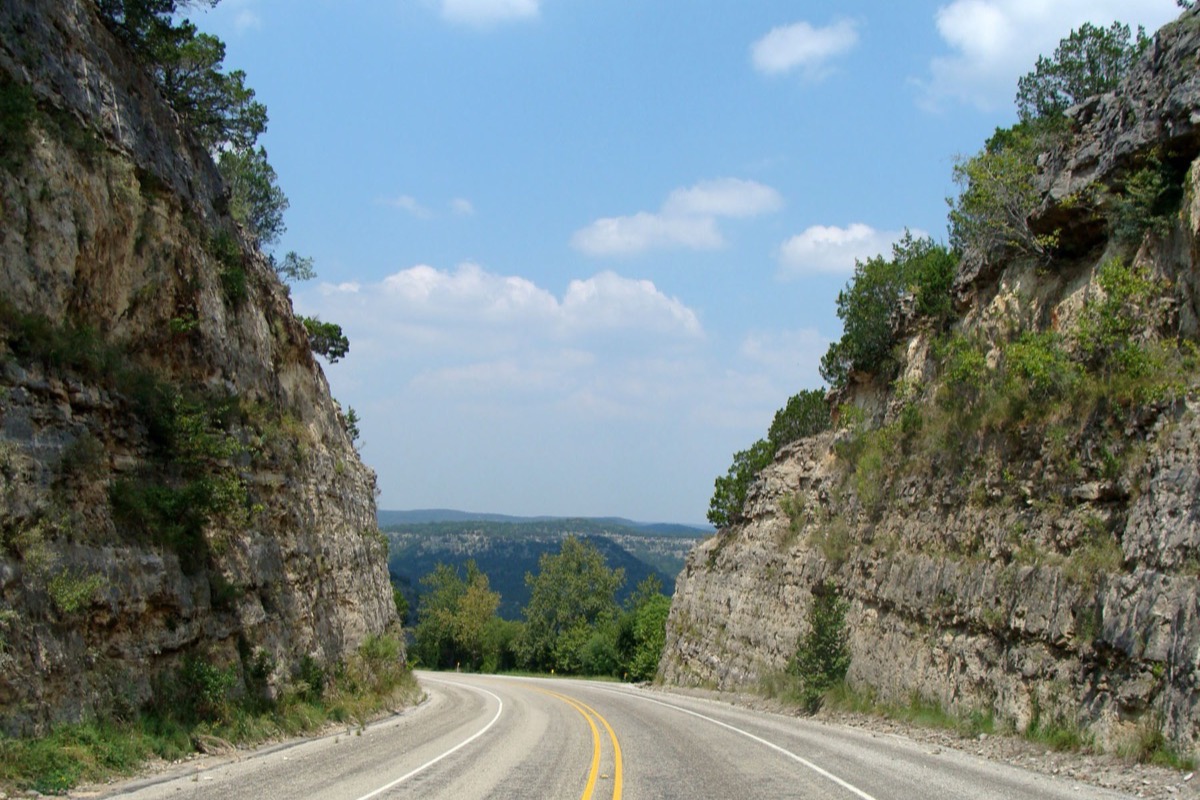 A Texas road curving between two high cuttings of layered limestone, scrub oak growing out of the ledges above and the green hill country opening out in the gap ahead.