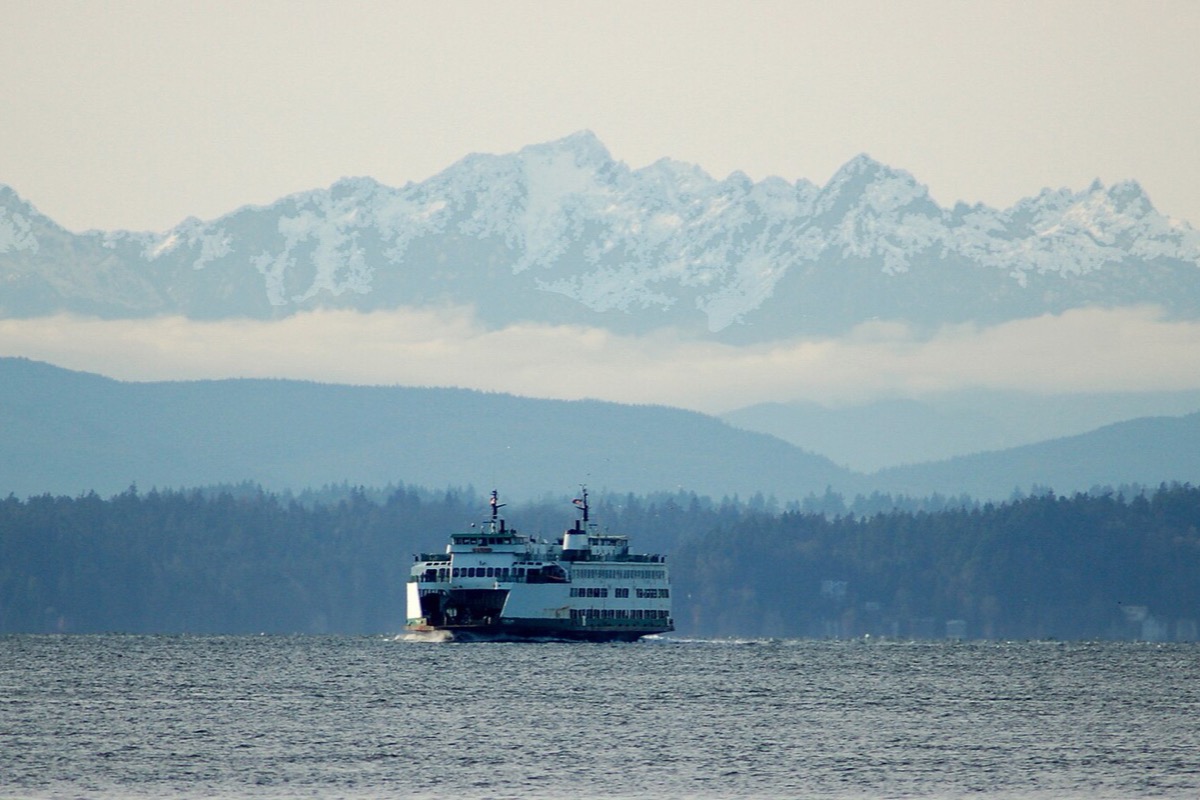 A Washington state ferry crossing flat grey water on Puget Sound, low forested islands behind it and the snow covered peaks of the Olympic range standing above the haze.