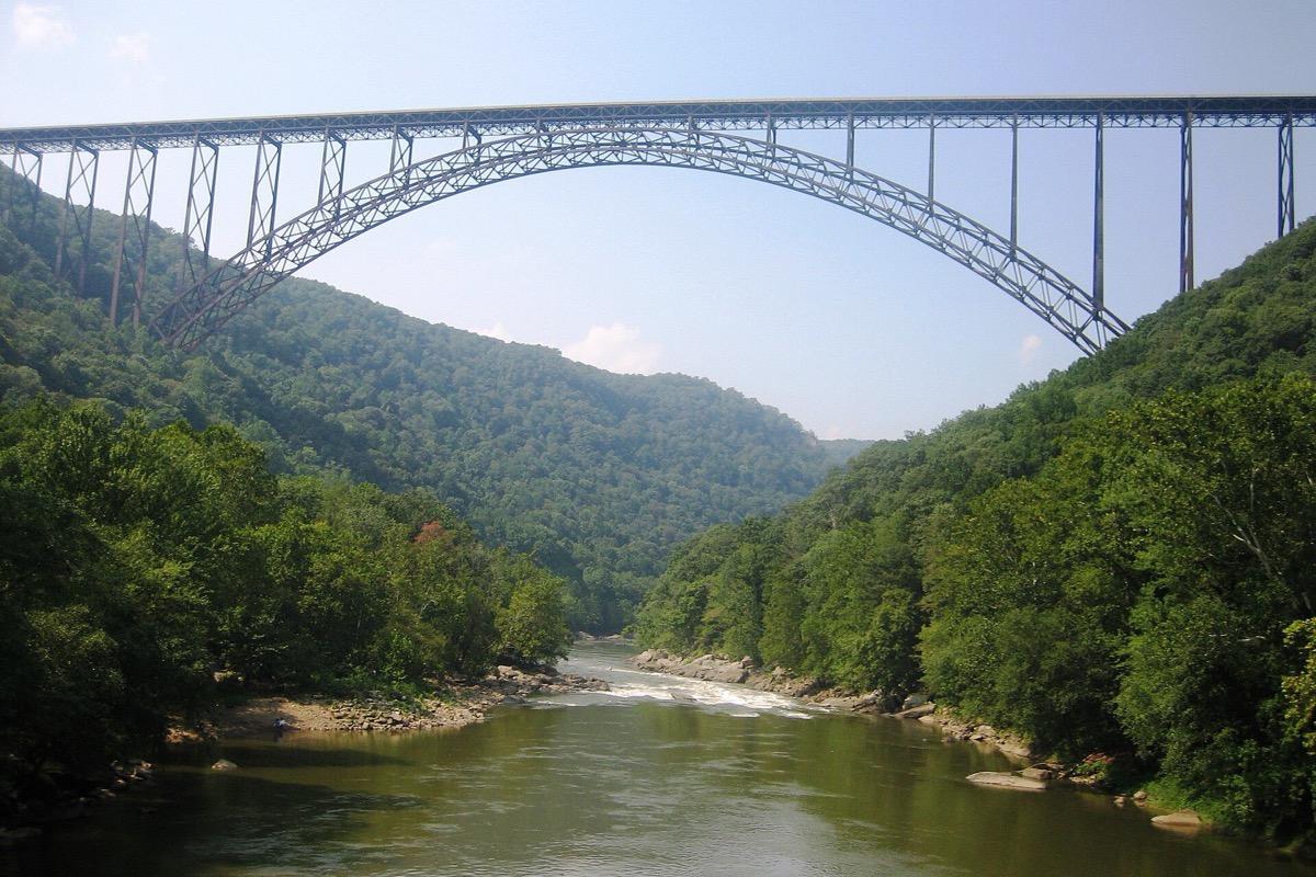 A single long steel arch carrying a West Virginia road high across a deep wooded gorge, the river running shallow over gravel bars far below and forested walls rising on both sides.