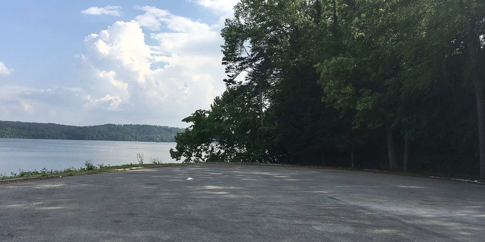 Empty paved lot beside a calm lake, tall green trees right, cumulus clouds overhead.