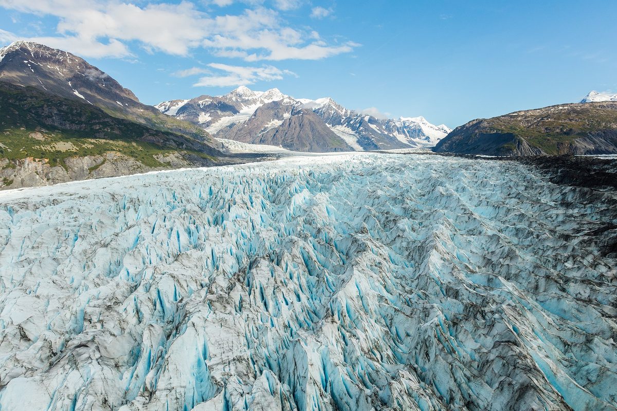 A crevassed Alaska glacier fills the valley in jagged blue ice ridges, running back toward snow-streaked peaks under a clear sky.