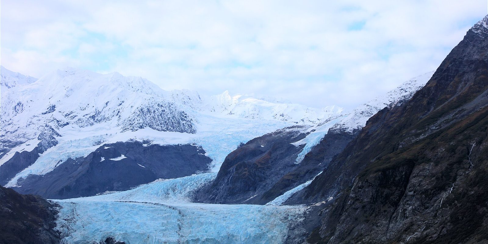 A pale blue glacier spilling between dark rocky slopes below snow-covered peaks under a cloudy sky