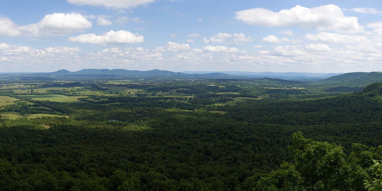 Wooded hills falling away to farm fields and low distant ridges under scattered clouds