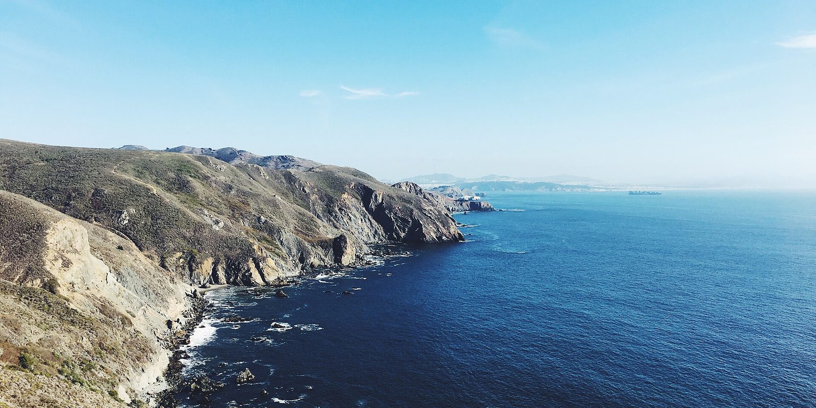 Dry coastal bluffs dropping to rocky surf, with hazy headlands along the far shoreline