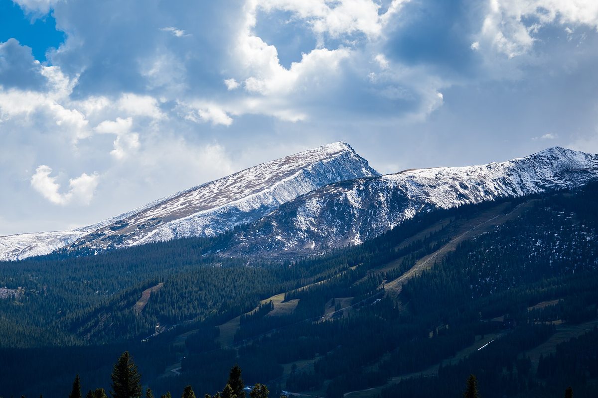 Snow-streaked Rocky Mountain peak rising above dark spruce forest and cleared ski runs in Colorado, with tall cumulus clouds behind it.