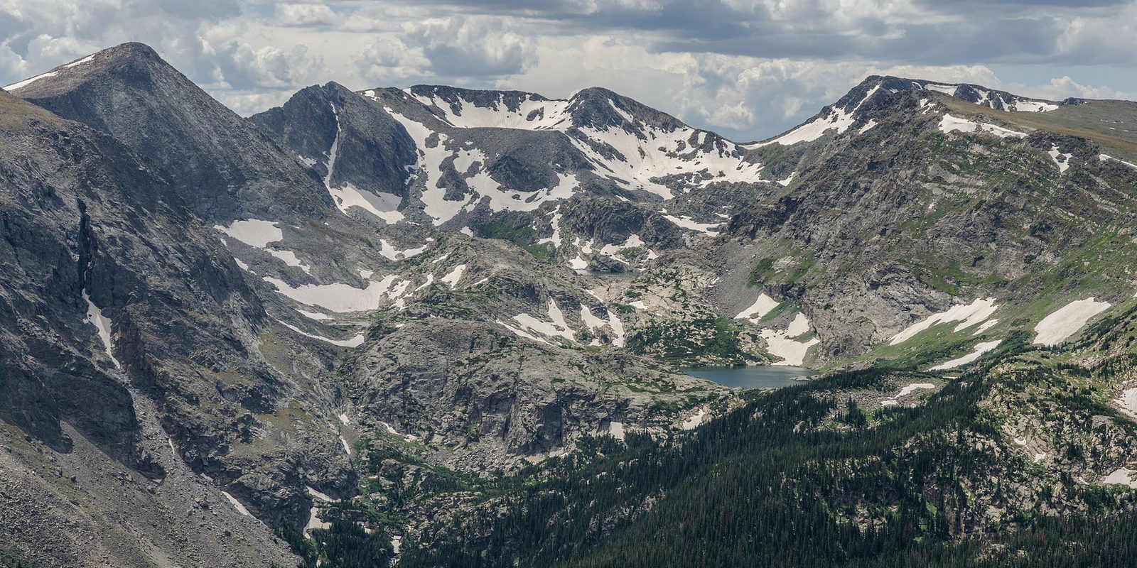 Rocky peaks streaked with summer snow above a small alpine lake and dark conifer forest