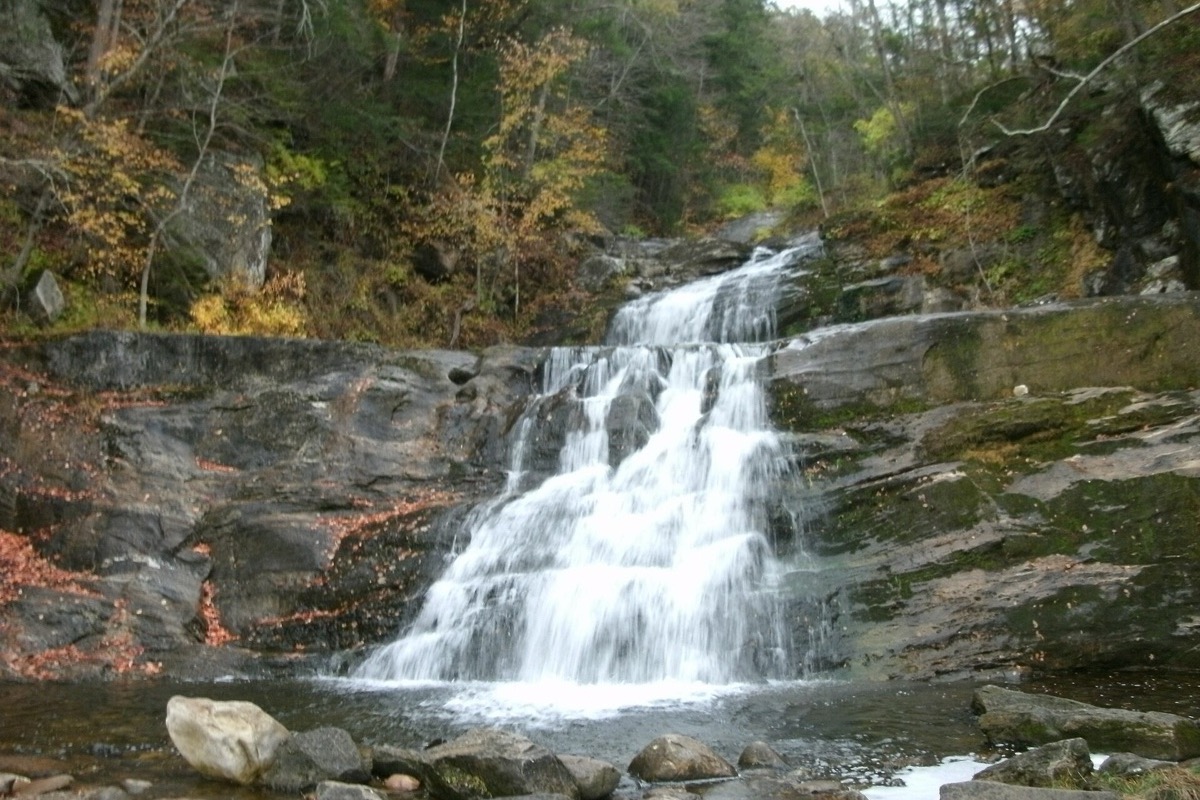 A Connecticut brook falling in white steps down a wide ledge of dark rock, autumn colour in the trees above it and pale boulders in the pool at the foot.