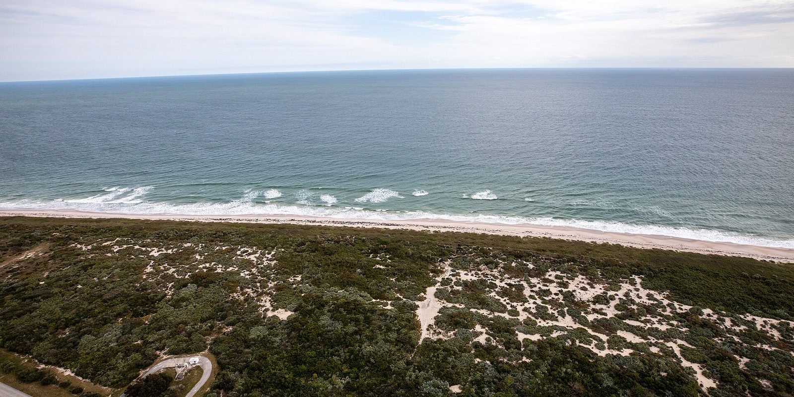 Aerial look over low coastal scrub and sand patches to a narrow beach and calm ocean