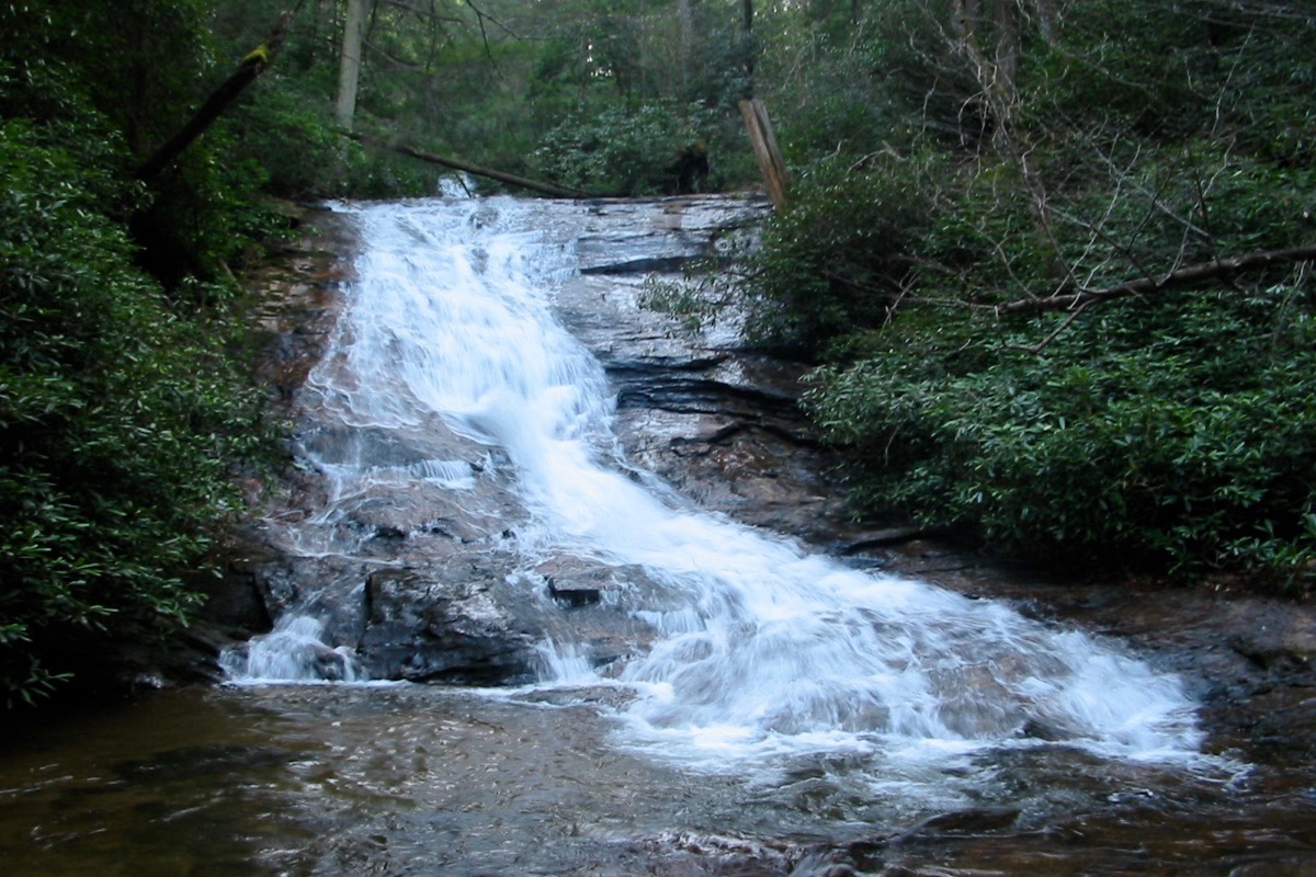 A north Georgia creek dropping in a long white cascade over a broad tilted rock face, dense green rhododendron and hardwood crowding both banks.