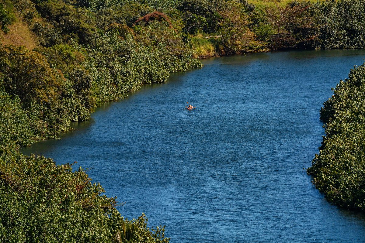 A lone kayaker paddling a wide blue river that winds between steep jungle-covered banks in Hawaii.