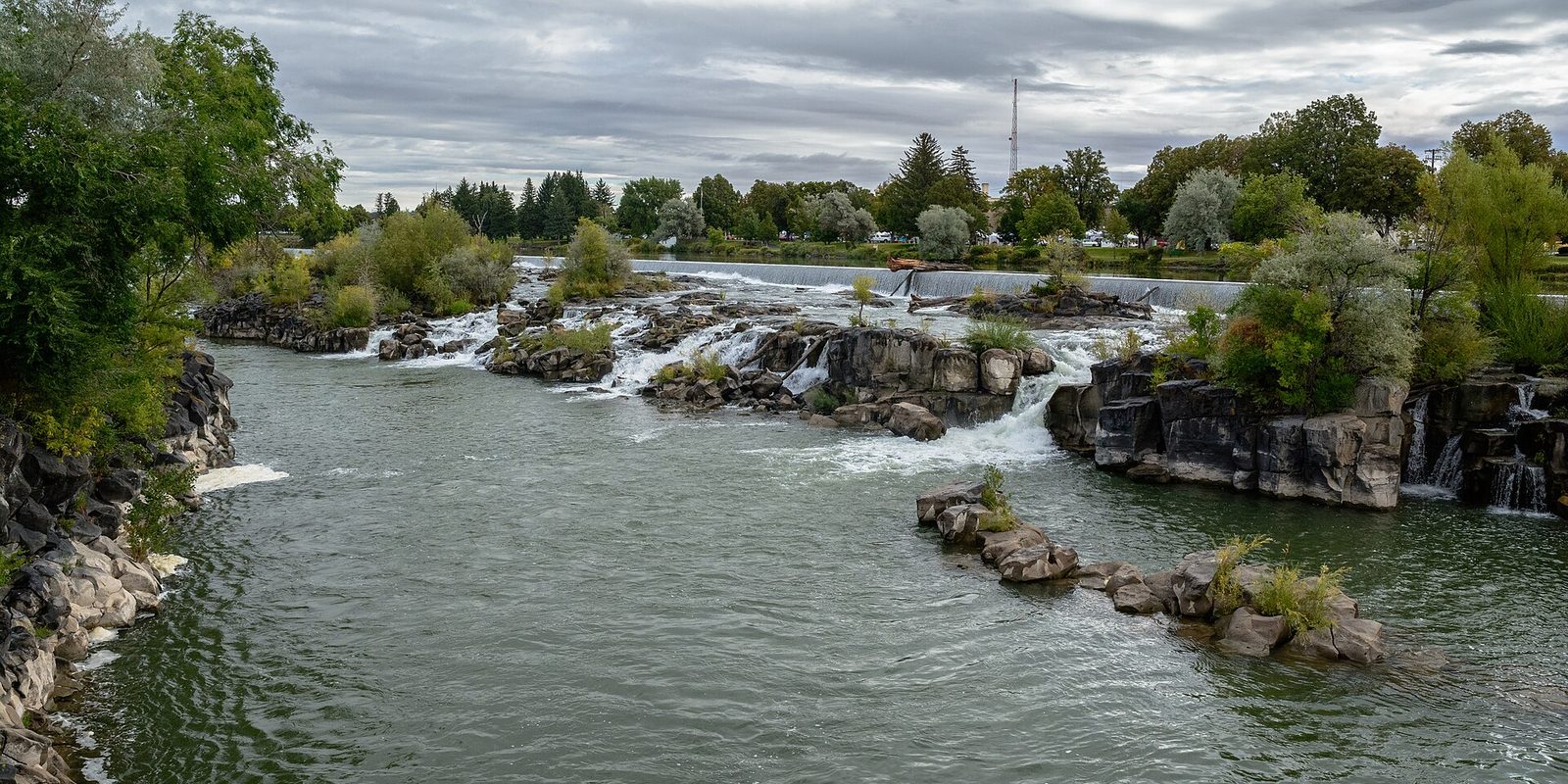 A broad river spilling over low rocky ledges below a treed park bank and grey clouds.